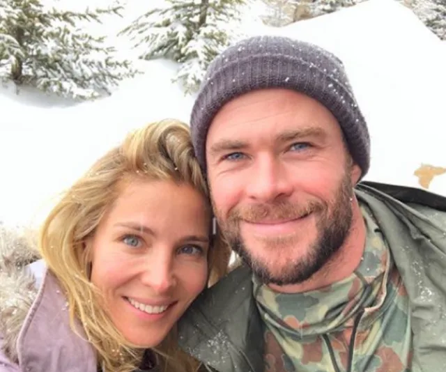 Couple in snowy landscape, man wearing a beanie and camouflage jacket, woman with long blonde hair, both smiling at the camera.