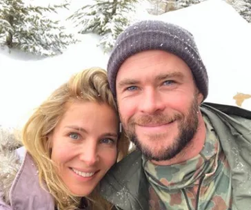 Couple in snowy landscape, man wearing a beanie and camouflage jacket, woman with long blonde hair, both smiling at the camera.