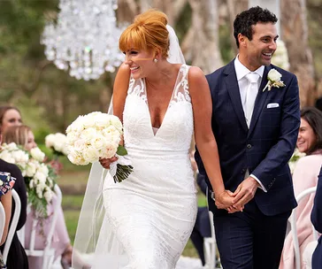 A bride and groom happily walk down the aisle, holding hands and smiling, surrounded by guests in an outdoor setting.