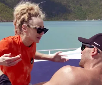 A woman in sunglasses talks emotionally to a shirtless man on a boat with a scenic water backdrop.