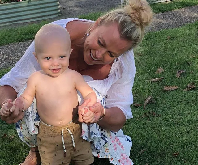 Woman kneeling on grass, joyfully holding a shirtless toddler's hands as he smiles and stands.