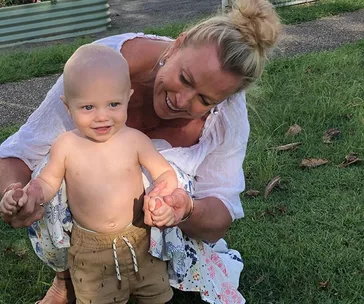Woman kneeling on grass, joyfully holding a shirtless toddler's hands as he smiles and stands.