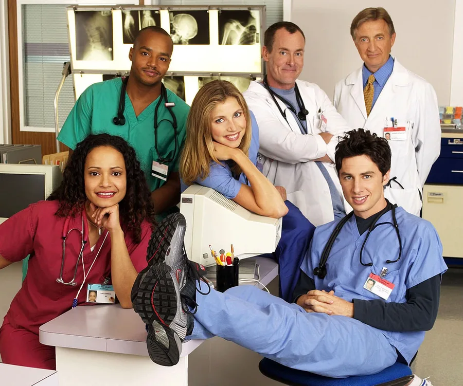 Cast of "Scrubs" in a hospital set, featuring characters in medical scrubs and lab coats, posing together smiling.