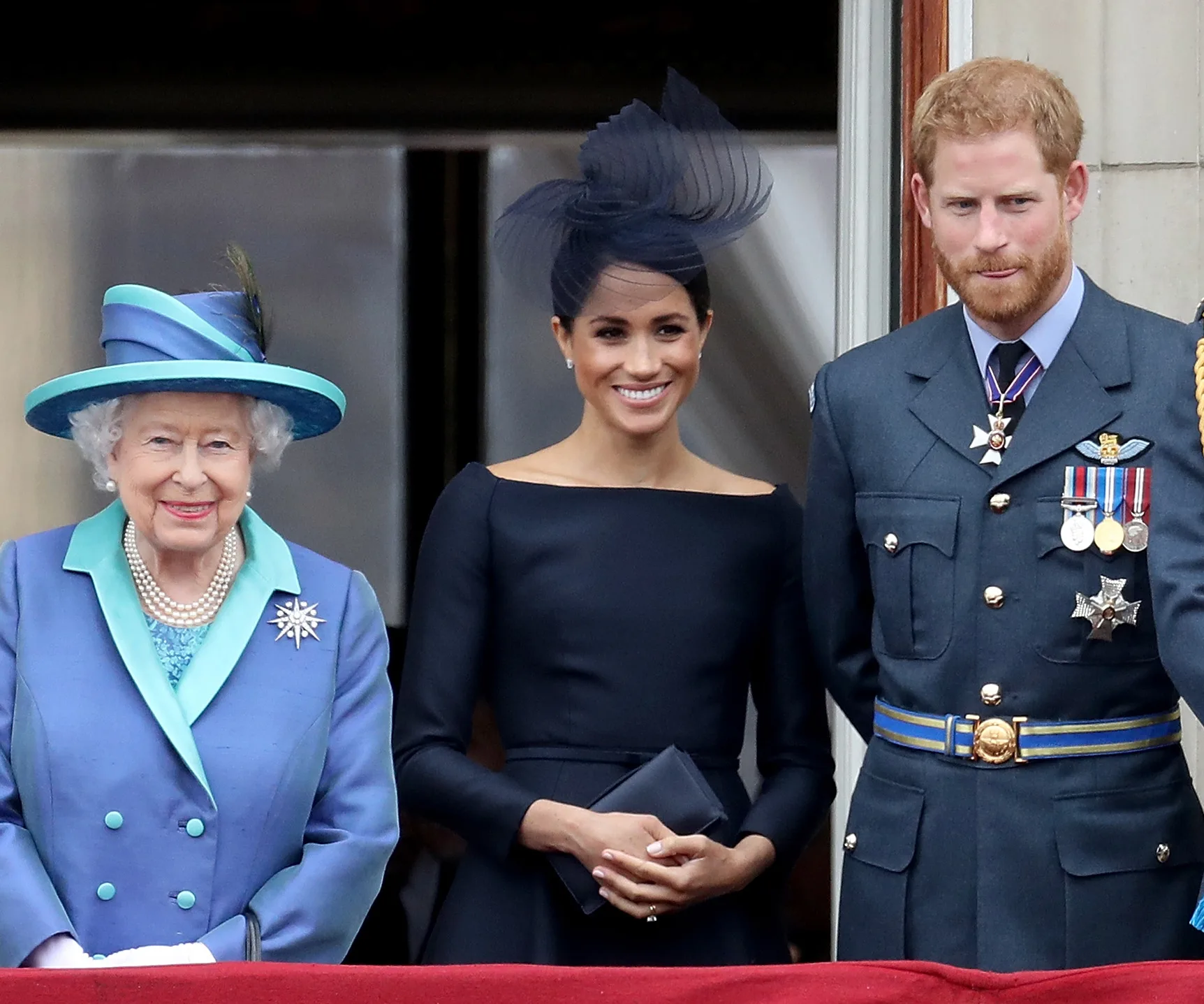 Queen Elizabeth II with Meghan Markle in a black dress and Prince Harry in military attire on a balcony.