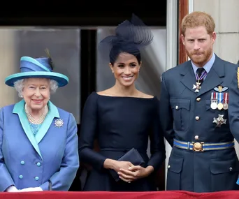 Queen Elizabeth II with Meghan Markle in a black dress and Prince Harry in military attire on a balcony.