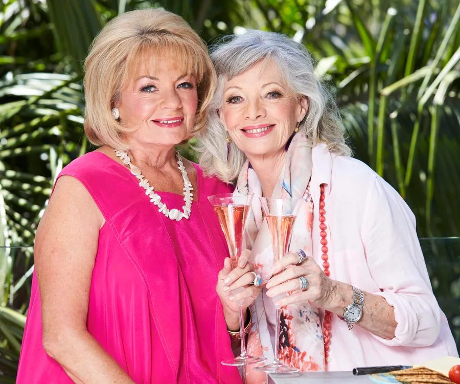 Two women smiling and holding champagne glasses outside, one in pink and the other in white with a scarf and jewelry.