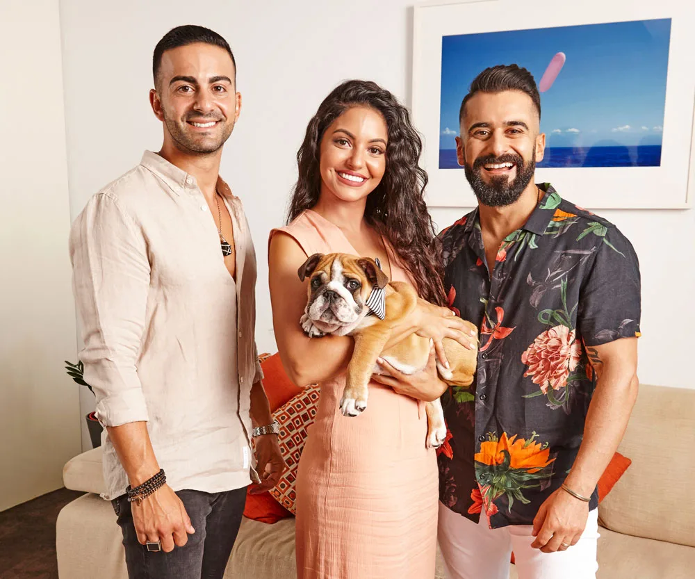 A woman holds a bulldog, standing between two men in a living room, smiling at the camera.