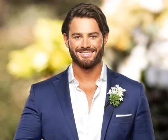A man in a blue suit with a boutonniere, smiling against a blurred outdoor background.