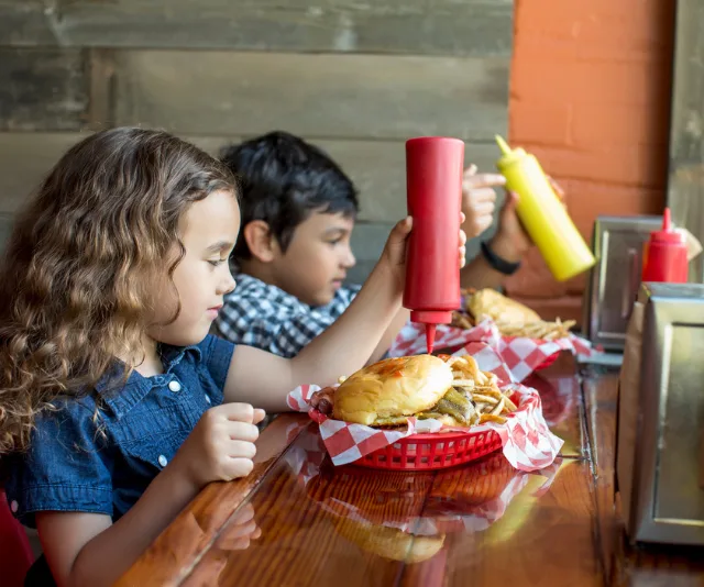 Children adding ketchup and mustard to burgers at a restaurant table.