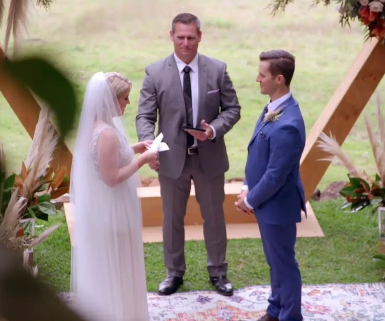 A couple stands at an outdoor wedding altar with a celebrant, exchanging vows.