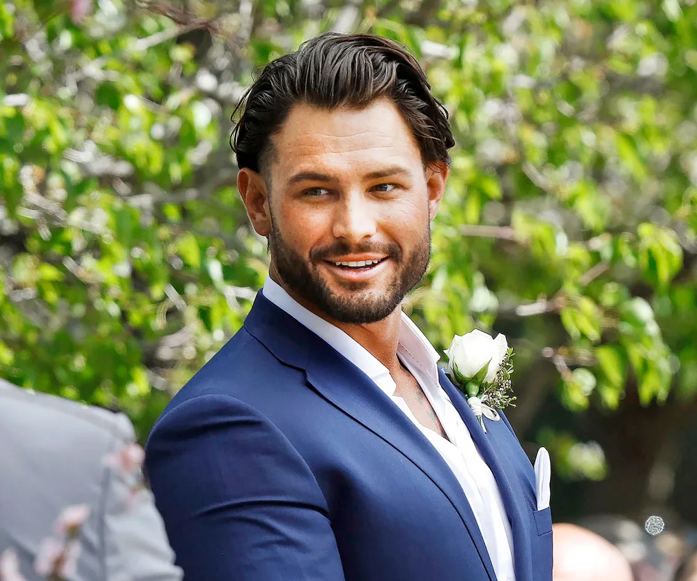 A man in a blue suit smiles outdoors, wearing a white rose boutonniere.