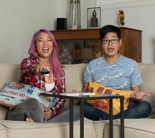 Two people sitting on a sofa, holding pillows, laughing and talking, one holding a remote in a cozy living room.