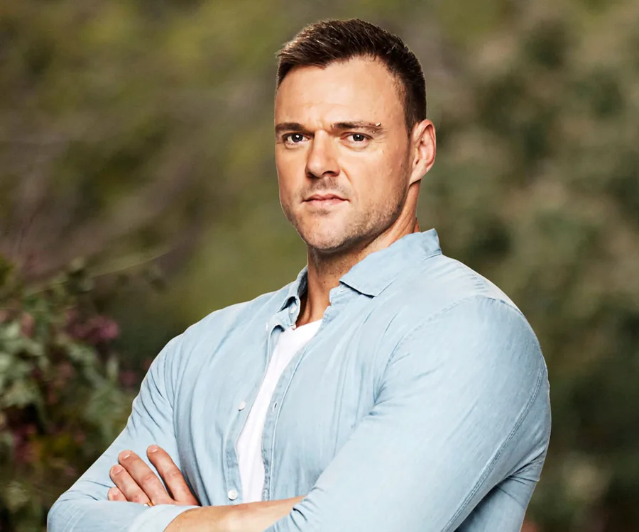 Man in light blue shirt with arms crossed, standing outdoors against a blurred green background.