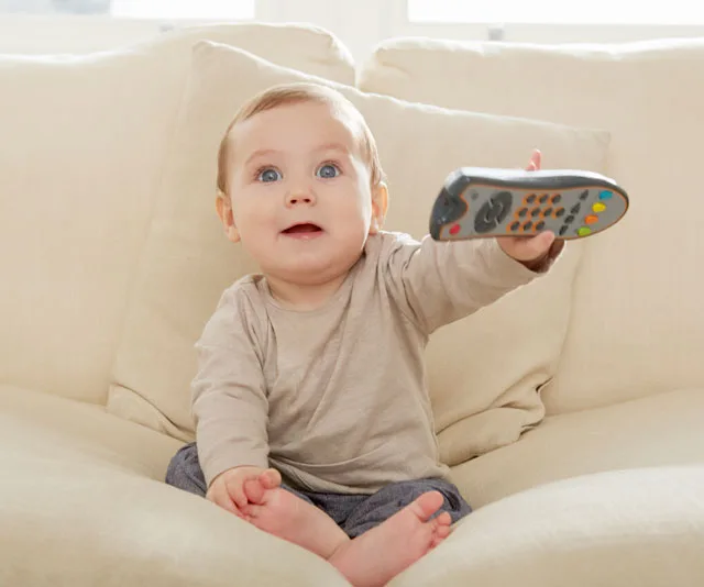 Baby sitting on a sofa holding a TV remote, looking up with an intrigued expression.
