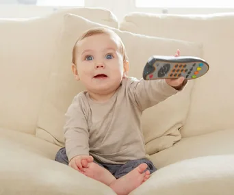 Baby sitting on a sofa holding a TV remote, looking up with an intrigued expression.