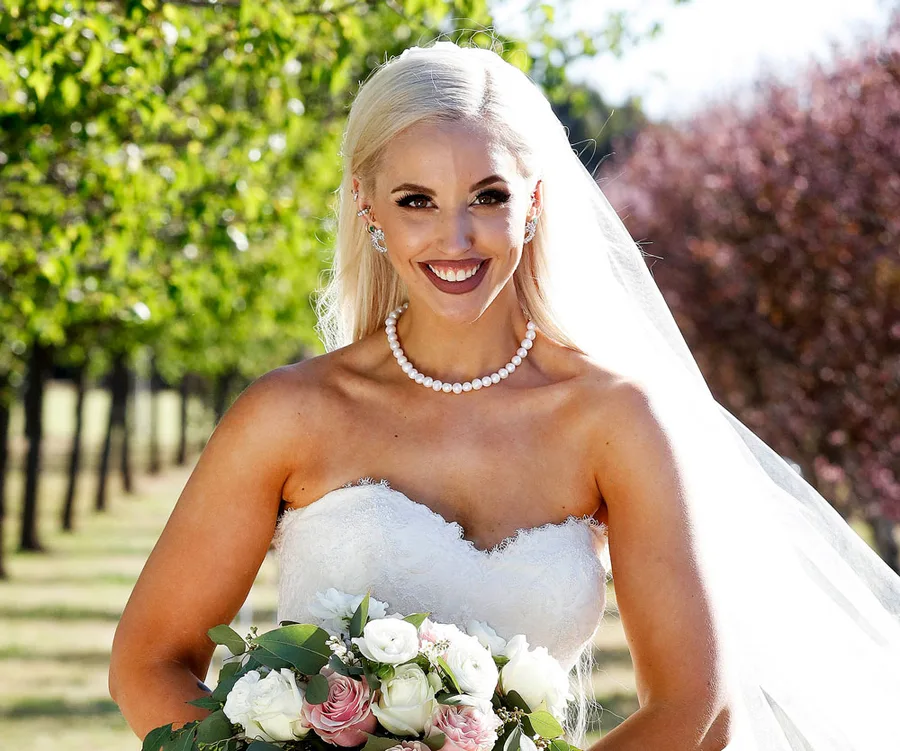 A bride in a white strapless dress holding a bouquet, smiling outdoors with trees in the background.