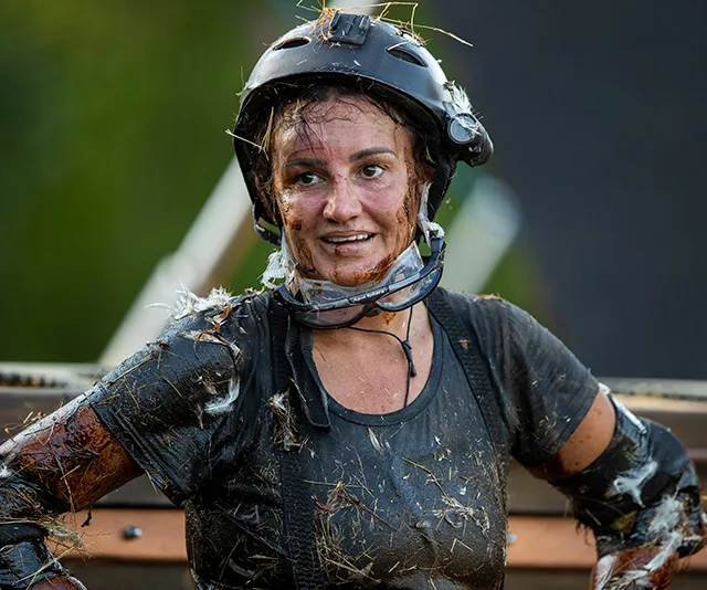 Jacqui Lambie covered in mud and straw, wearing a helmet and gear, appears tired but determined on "I'm a Celebrity."