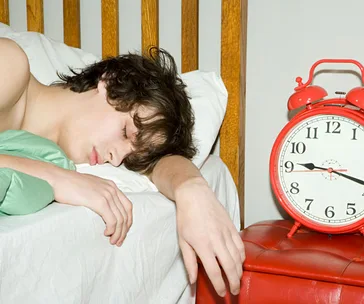 Teen asleep in bed next to a red alarm clock showing 7:10.