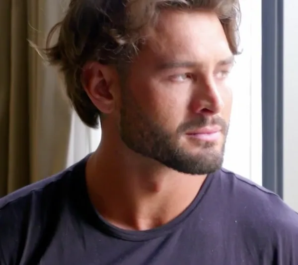 Bearded man gazing out of a window in thought, wearing a dark shirt, with natural light illuminating his face.