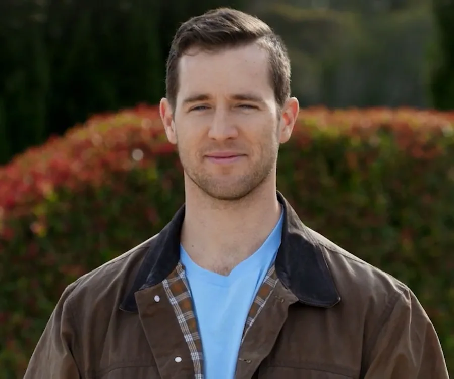Man wearing a brown jacket over a light blue shirt, standing outdoors with blurred greenery in the background.