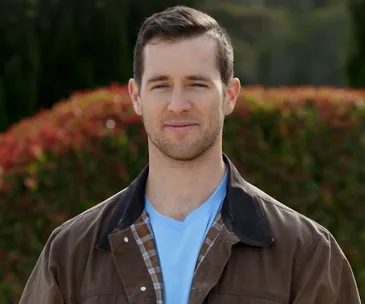 Man wearing a brown jacket over a light blue shirt, standing outdoors with blurred greenery in the background.