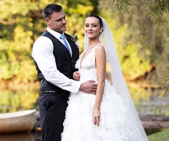 A bride and groom pose by a serene lake, with the groom in a suit and the bride in a floral wedding dress and veil.