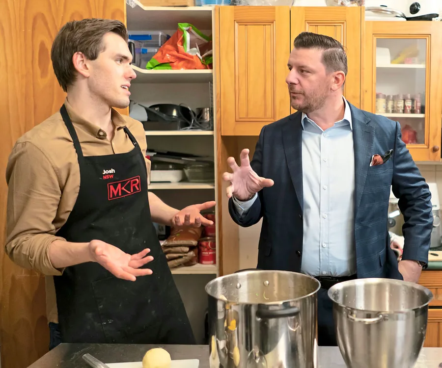Josh in an apron listens to a man gesturing in a kitchen during "My Kitchen Rules" episode discussion.