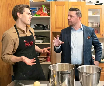 Josh in an apron listens to a man gesturing in a kitchen during "My Kitchen Rules" episode discussion.