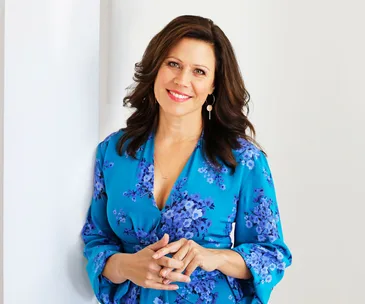 A woman in a blue floral dress, smiling, with long brown hair, standing against a white background.