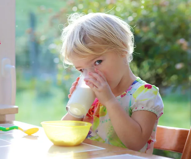 A young child with blonde hair drinks milk from a glass at a table with a yellow bowl and spoon.