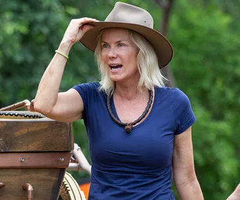 Woman in blue shirt and wide-brimmed hat, outdoors, gesturing near wooden cart.