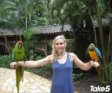 A person smiling, holding two colorful parrots on outstretched arms, with a tropical backdrop and "Take 5" logo in corner.