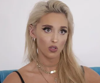 ```plaintext
Woman with blonde hair and dramatic makeup looking surprised, wearing layered necklaces and multiple earrings.
```
