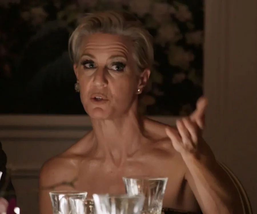 A person with short hair gestures while speaking at a dinner table, glassware visible in the foreground.