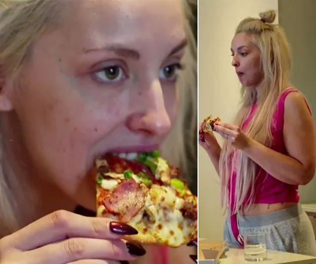 A woman with long blonde hair eating a slice of pizza, wearing a pink top and gray pants, in a casual home setting.