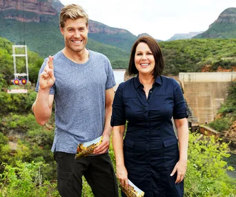 Two people smiling outdoors, holding "I'm a Celebrity" cards, with greenery and a dam in the background.