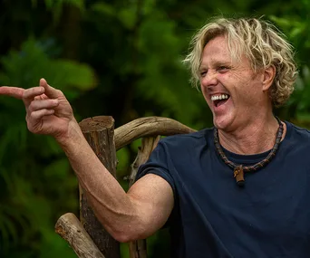 Man in a jungle setting, laughing and pointing with an animated expression while wearing a dark t-shirt and necklace.
