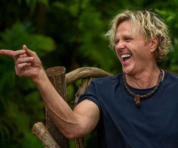 Man in a jungle setting, laughing and pointing with an animated expression while wearing a dark t-shirt and necklace.