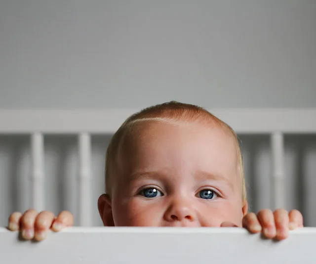 Baby peeking over crib railing, showing only eyes and forehead, in a dimly lit room.