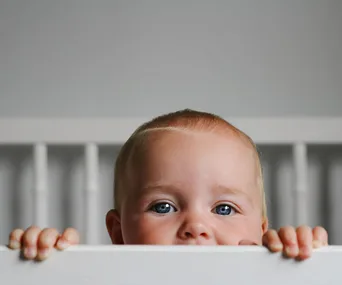 Baby peeking over crib railing, showing only eyes and forehead, in a dimly lit room.