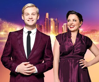 Two hosts in formal attire, smiling against a vibrant cityscape background at Eurovision Song Contest 2019.