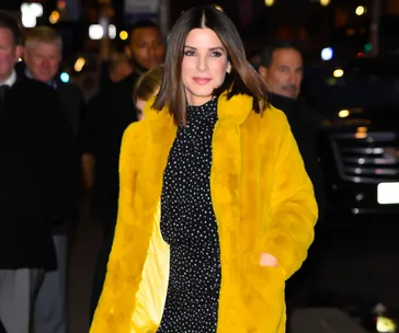 Sandra Bullock in a polka dot dress and bright yellow coat walking on a city street at night.