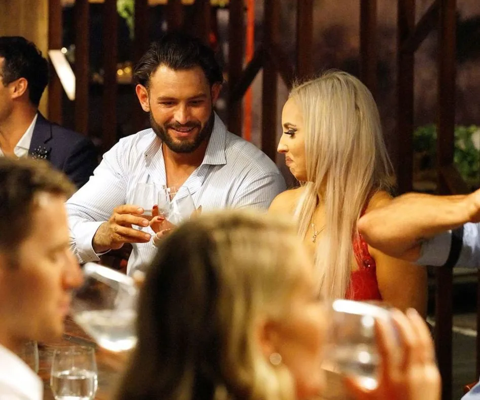 A man and a woman are seated at a dinner table, holding glasses, engaged in conversation with others around them.
