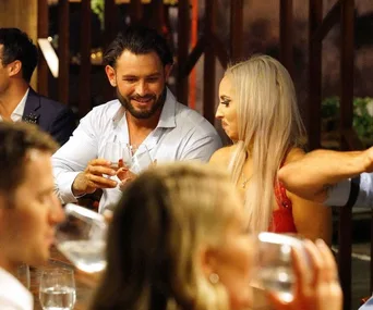 A man and a woman are seated at a dinner table, holding glasses, engaged in conversation with others around them.