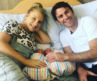 A smiling couple lies in bed holding their newborn twin babies wrapped in striped blankets.