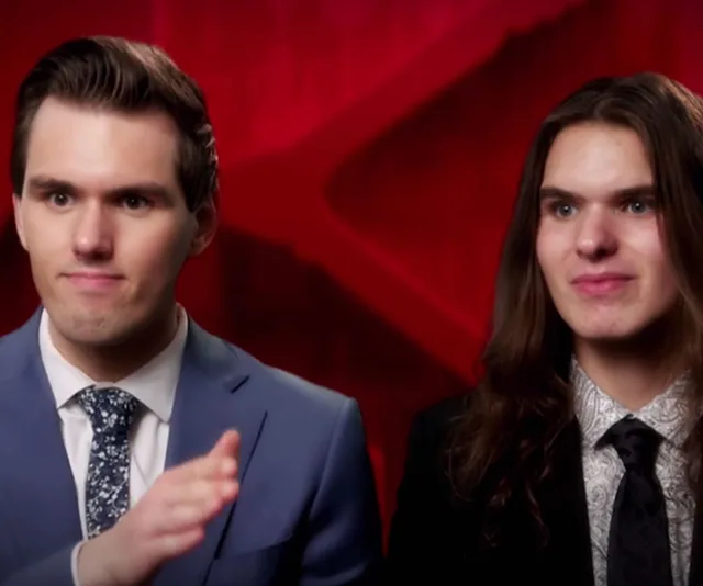 Two men in suits speaking in front of a red background on a television set.