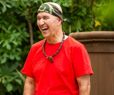 Tahir Bilgic in a red shirt and camo headband, smiling outdoors on "I'm a Celebrity".