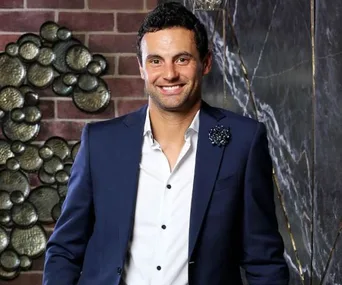 Man in a navy suit with a floral lapel pin smiles against a brick and marble wall background.