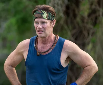 Man in a blue tank top and camouflage headband standing outdoors with hands on hips.