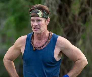 Man in a blue tank top and camouflage headband standing outdoors with hands on hips.